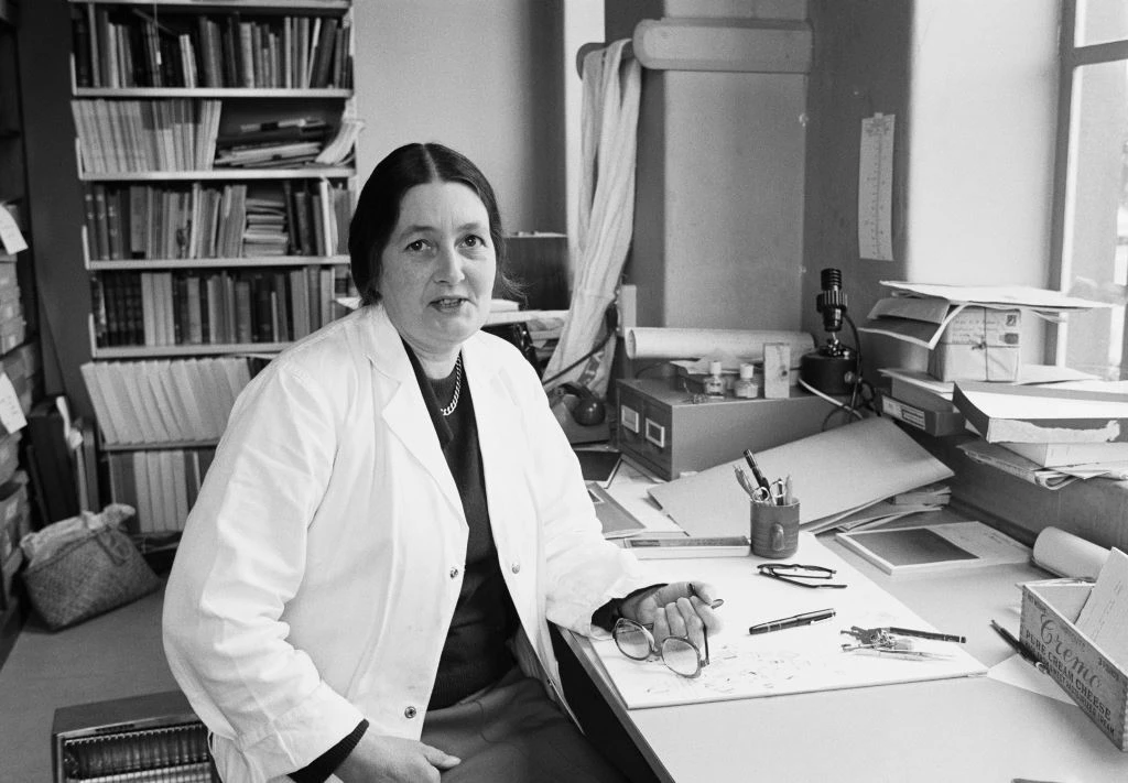 A black and white photo of a woman in a white coat sitting at a desk and looking at the camera.