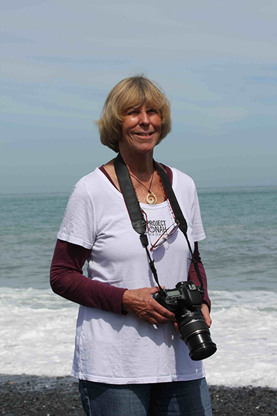 A woman holding a camera is standing in front of the sea.