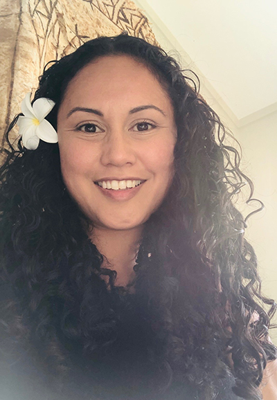 Head and shoulders of a woman with long hair and a frangipani flower over one ear. She is smiling at the camera.