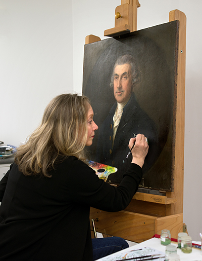 A woman is touching up an old painting on an easel in a studio.