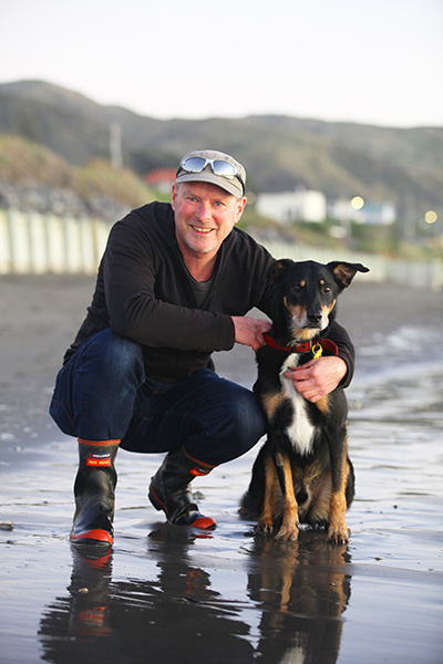 A man is crouched down next to his dog on a beach. The man is smiling at the camera.