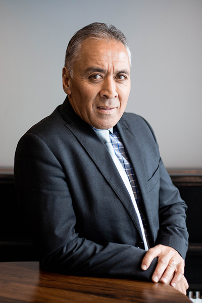 A photo of a man in a suit leaning one arm on a table and looking at the camera
