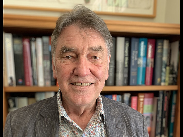 Peter Simpson Top half of a man standing in front of a bookshelf and smiling at the camera.