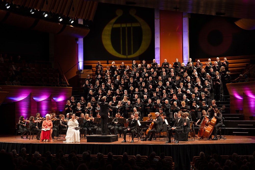 A large choir is assembled on a stage. There are musicians and promenant singers in front of them.