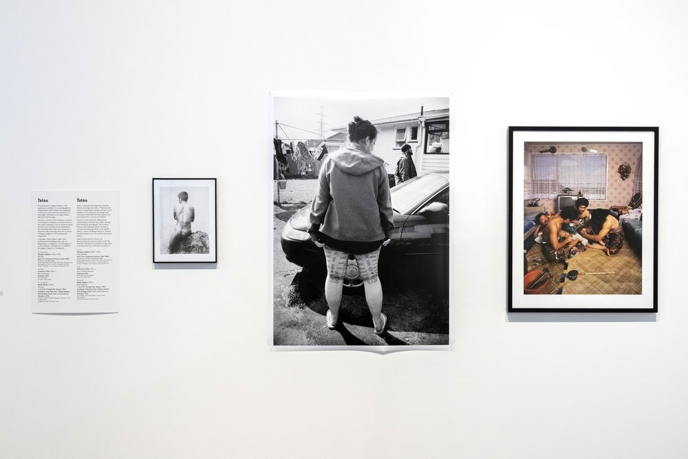 A photo of an exhibition wall with three photos on it of people either with traditional tattoos or receiving one.
