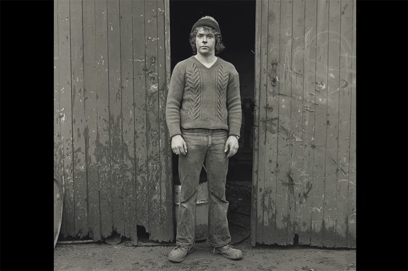 A black and white photo of a man standing in front of a shed.