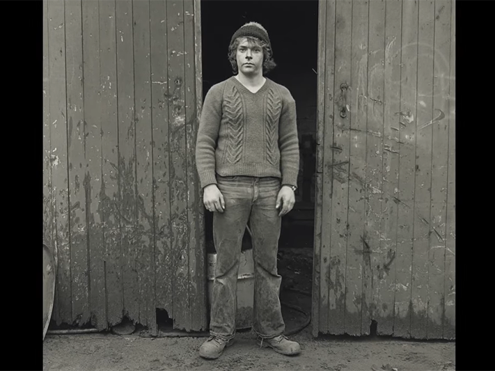 Peter Andrew King, moulder, cast iron foundry, Christchurch, 1981. From the series: Working Men A black and white photo of a man standing in front of a shed.