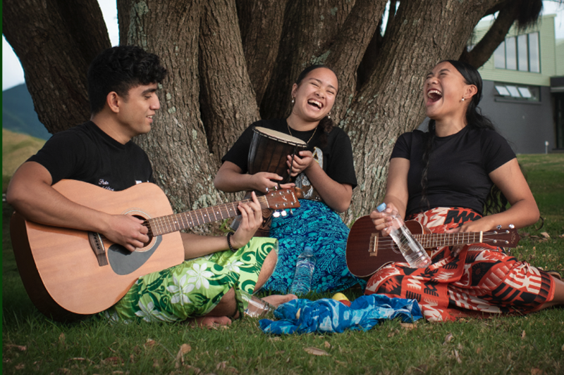 Three young people are sitting under a tree. One is playing the guitar, one is playing a drum and one has a ukulele. They are all laughing.