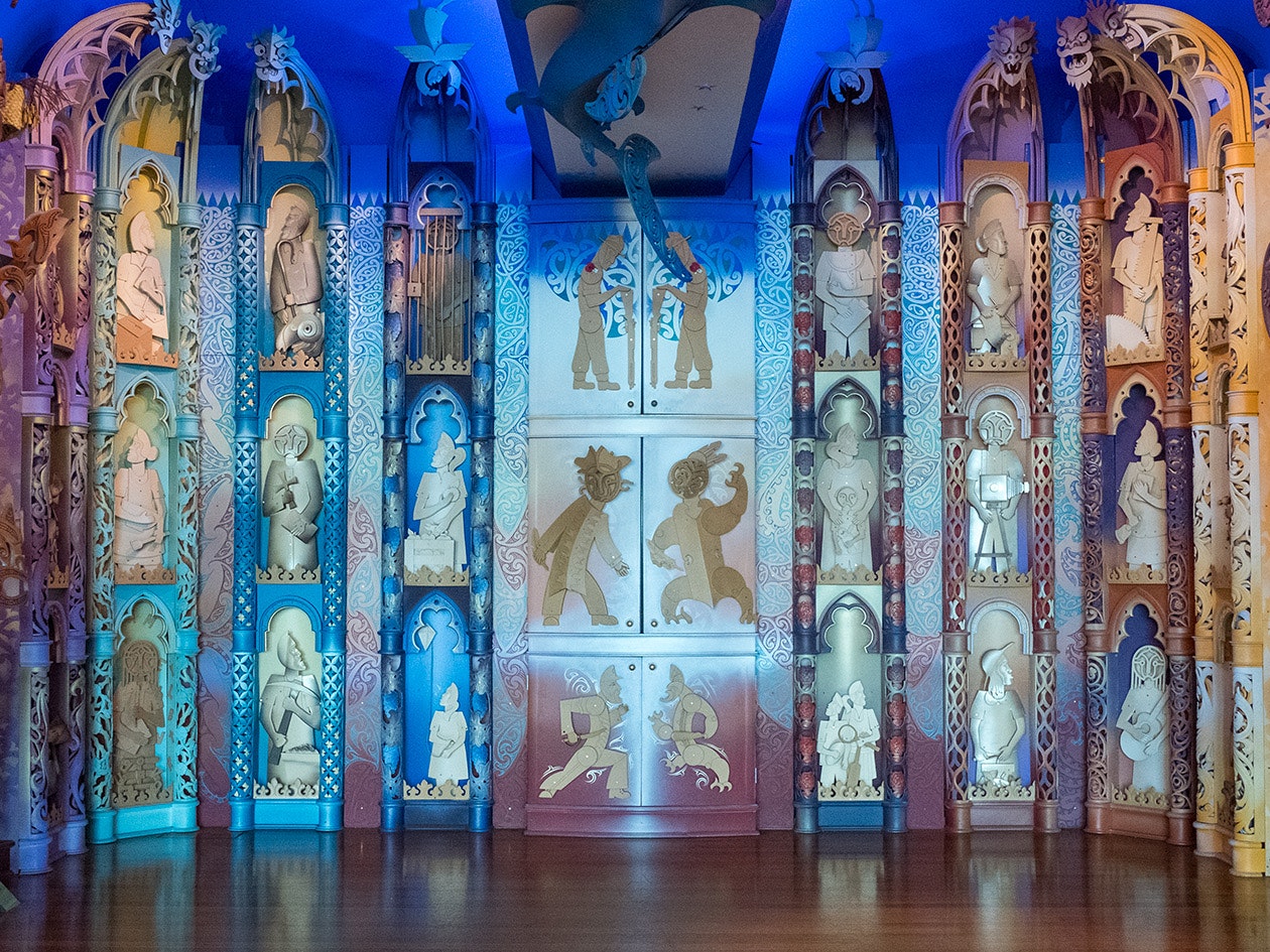 Detail of Te Hono ki Hawaiki, 2024. Photograph by Jane Harris. Te Papa A colourful wall with carvings of stylised people doing different things and three cupboards in the central section.