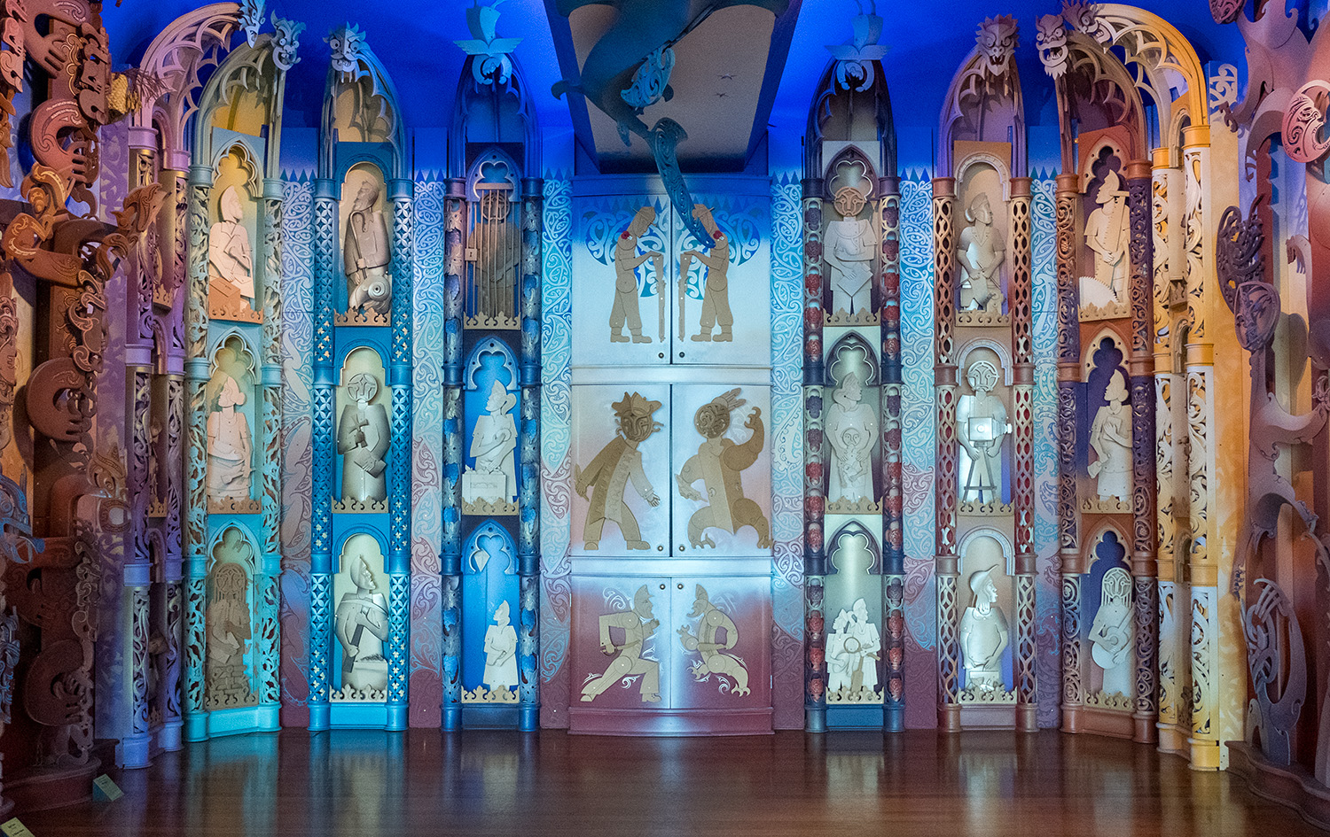 A colourful wall with carvings of stylised people doing different things and three cupboards in the central section.