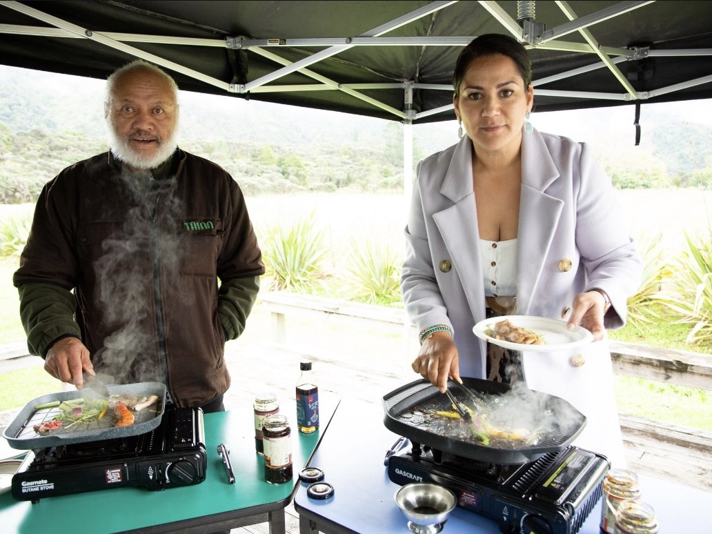 Rereata Makiha and Dallas King. Photo Courtesy of Dallas King Two people standing under an open marquee tent. Both are cooking on a frypan and  looking at the camera.