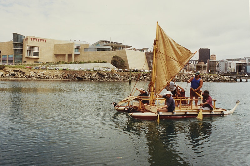 Five people are paddling a boat in front of a building with angular shapes.