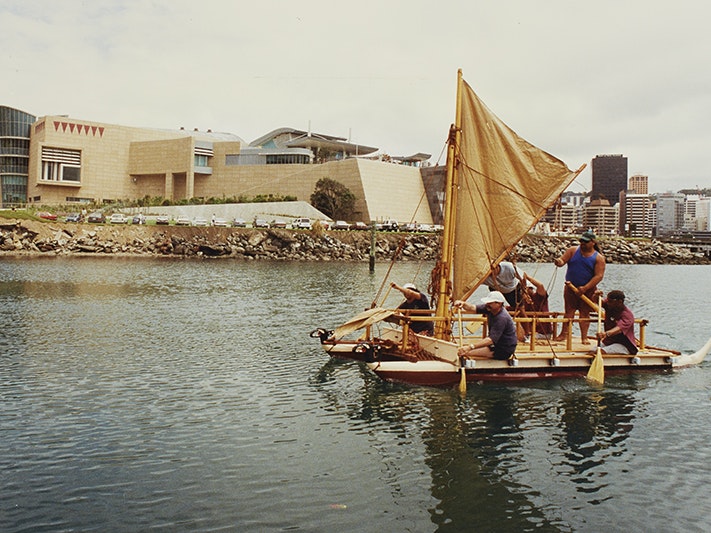 Te Papa workers and crew paddle Te Aurere Iti to the official opening ceremony of Te Papa in Wellington, 1997. Te Papa Five people are paddling a boat in front of a building with angular shapes.