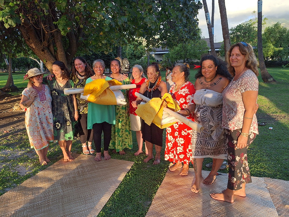 Several women standing in a group in the late afternoon sun. Some of them are holding bundles of tapa.