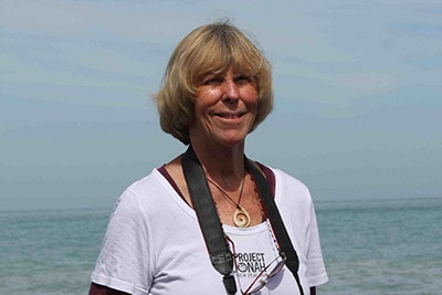 Head and shoulders of a woman smiling. The ocean is in the background.