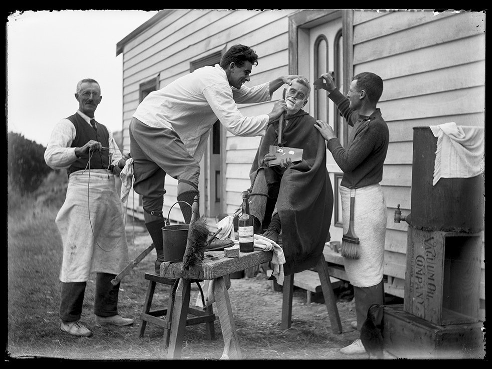 Leslie Adkin, Preparations for a return to civilization, 28 February 1921, Kāpiti Island. Gift of G. L. Adkin family estate, 1964. Te Papa (B.022218) A black and white photo of four men outside a house. One of them is sitting in a tall chair and being shaved by another man.