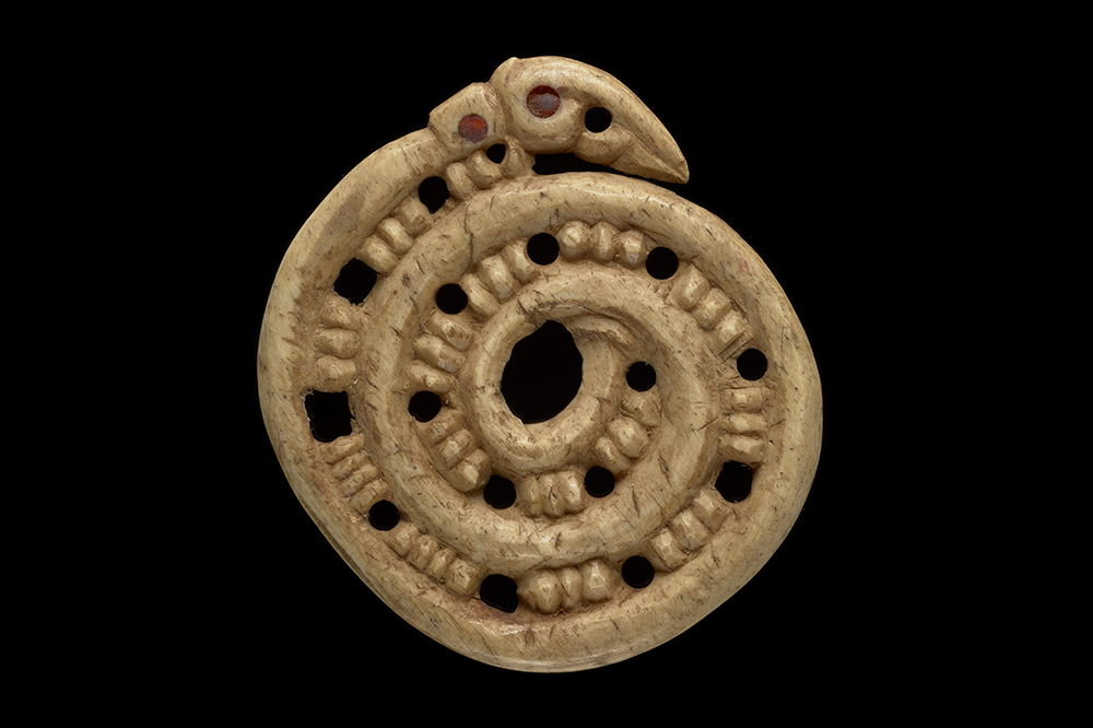 A spiral carving with holes every four notches. There is a ahead at the end of the spiral with red beads for eyes. He whakairo tōrino whai kōhao i ia whā pākati. Kei te pito o te tōrino tētahi upoko whai pirepire whero hei karu.