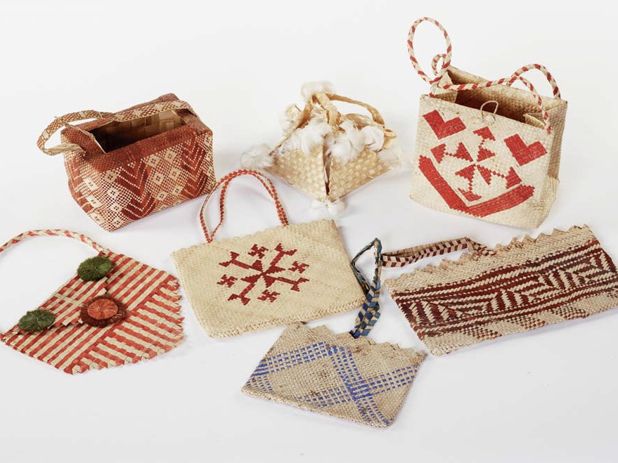 Left to right: kete p.p. (satchel) Cook Islands, date unknown, pandanus fibre, 355 x 140mm (Te Papa FE012041); kete po’o (basket), Cook Islands, date unknown, pandanus fibre, 890 x100mm (Te Papa FE012031); kete p.p., Cook Islands, date unknown, pandanus fibre, 210 x240mm (Te Papa FE012034); kete p.p. kaka‘o, Cook Islands, date unknown, fernland reed (Miscanthus floridulus), 120 x220 x 220mm (Te Papa FE012057); kete (basket), Cook Islands, date unknown, pandanus fibre, 200x130mm (Te Papa FE012032); kete p.p., Cook Islands, date unknown, pandanus fibre, 195 x320mm (Te Papa FE012035); kete p.p., Cook Islands, date unknown, pandanus fibre, 160 x 200mm (Te Papa FE012033). Artists unknown. Gift from Mr J.D.Hutchin, 1948. Seven woven items made of light brown, red, and blue pandanus leaves.