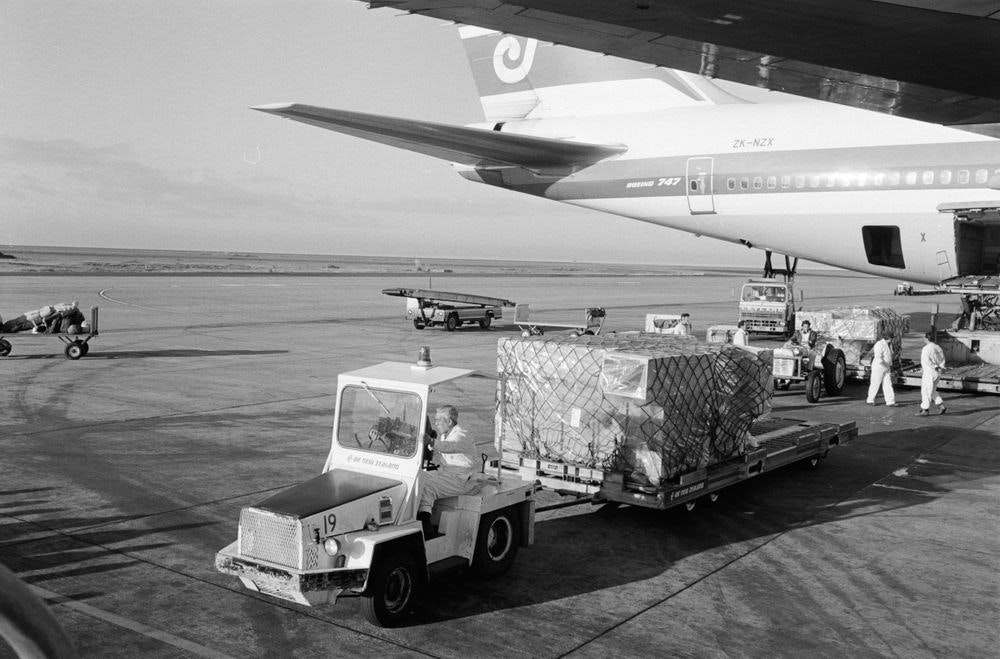 Te Māori exhibition, Brian Brake; photographer; about 1986; New Zealand A black and white photo of an airport runway with part of a plane and a luggage transport cart and trailer in the foreground