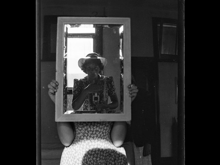 Self portrait, circa 1940s, maker unknown. Purchased 1999 with New Zealand Lottery Grants Board funds. Te Papa (B.080575) A black and white photograph of a woman sitting in a chair holding up a mirror which is reflecting another woman in a hat taking a photograph