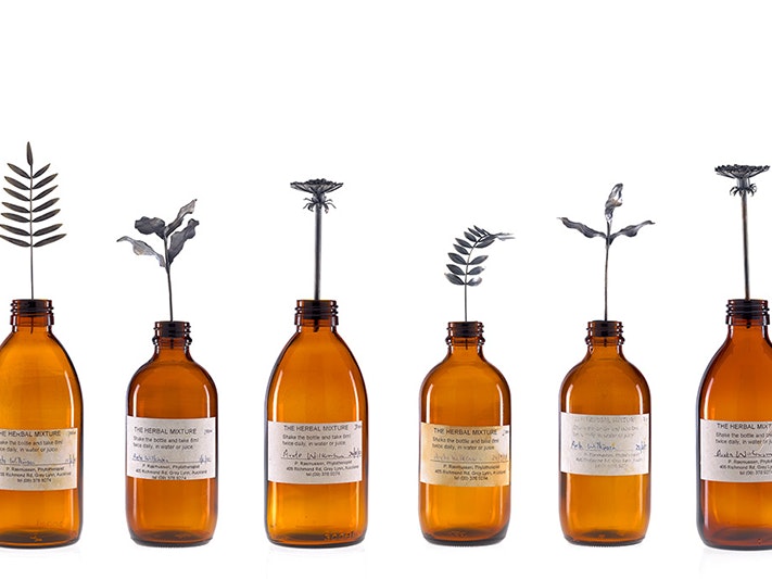 Crop of Herbal mixture II, 2001, by Areta Wilkinson (Ngāi Tahu,Kāti Māmoe, Waitaha) . Te Papa (2001-0039-1) A photograph of ten brown bottles with labels lined up, Each bottle has a single silver leaf attached to a stem sticking out of it.