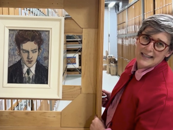 Still from The mysterious gaze of Edward McKnight Kauffer. Te Papa (243235) A woman in a red jacket is talking about a painting that is in an art store room. She has pulled it out on its rack.
