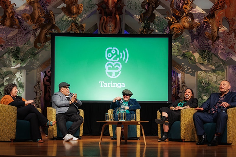 Taringa Live Podcast: Te Maori, 2024. Photo by Jo Moore. Te Papa (247569) Five people are sitting on a stage in a half circle. There is a large green screen behind them with a stylised design of a hei tiki.