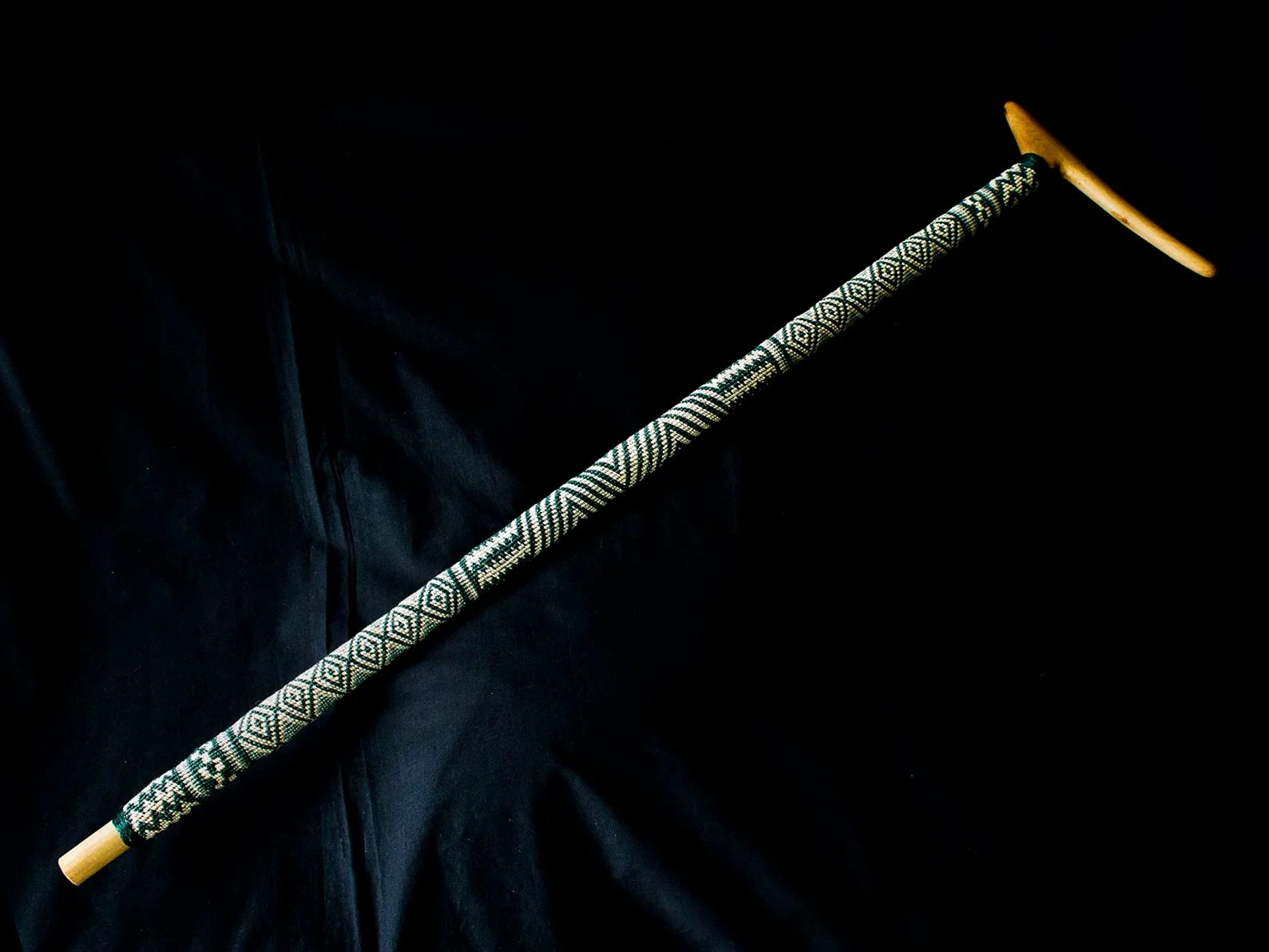 Tokotoko tāniko, woven by Adrienne Spratt, muka (flax fibre), dye, carved by Mitchell (Midi) Tareha, Kānuka, 2020. Te Papa (ME024646) A walking stick with a woven cover on the shaft.