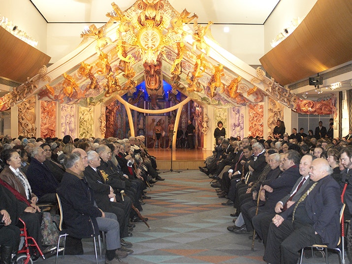 Mō Tātou: The Ngāi Tahu Whānui exhibition opening, 2006. Photo by Te Papa Imaging. Te Papa (70238) A large indoor spacec with a lot of people sitting in rows facing each other. At one end there is a carved and colourful stage.