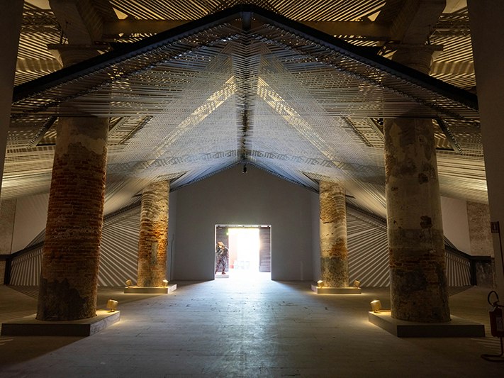 Takapau, 2022 at Stranieri Ovunque – Foreigners Everywhere, The 60th International Art Exhibition – La Biennale di Venezia. Photo by Ben Stewart A gallery with six pillars and an artwork strung up using wide material straps.