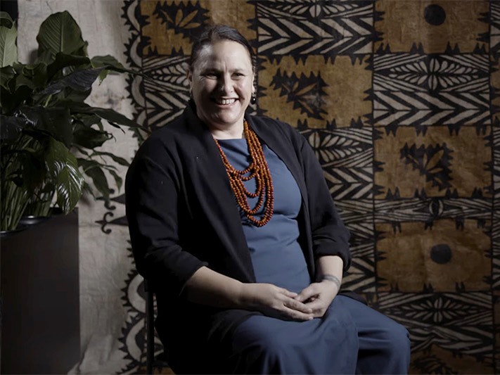 Liana Leiataua A woman is in a blue outfit and sitting in front of a large tapa cloth and smiling at the camera.