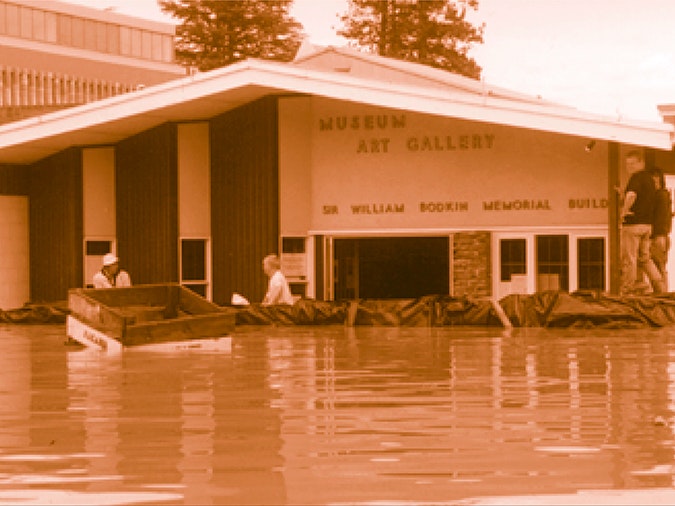 The photo is courtesy of Alexandra Museum A sepia toned photo of people retrieving items from a flooded museum. The water outside the building is waist high.