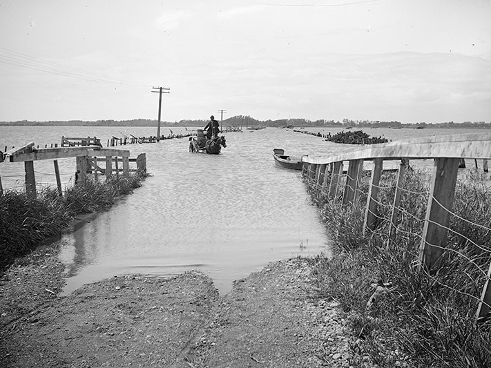 Flooded river, by Leslie Adkin. Gift of G. L. Adkin family estate, 1964. Te Papa (B.021624) A black and white photo of a man on a horse and cart crossing a flooded roadway.
