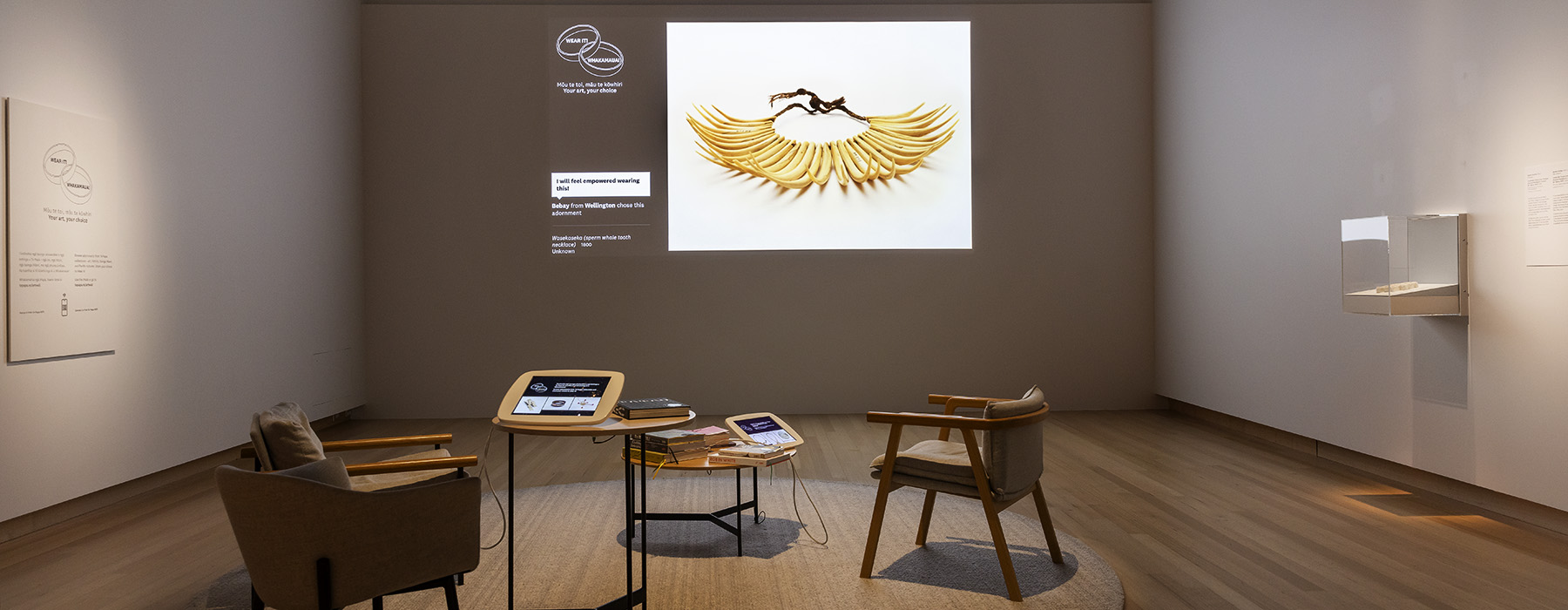 A photo of a space with chairs and a table, and a projection of a piece of jeweelery on the back wall.