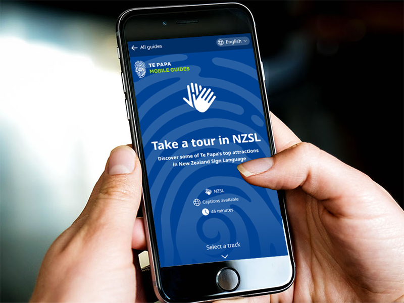 Te Papa NZSL mobile guide. Te Papa Closeup of hands holding a mobile phone which is displaying the NZSL mobile guide.