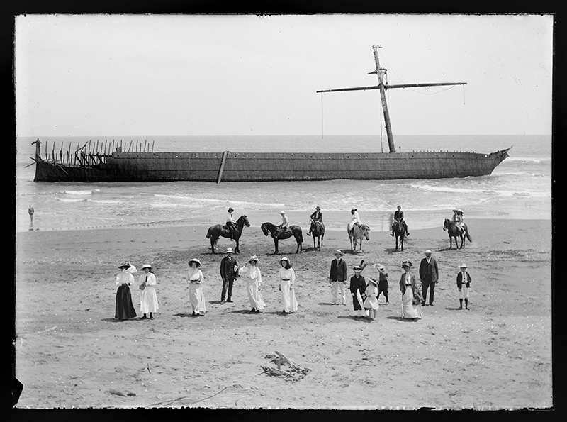 A black and white photo of about 18 people on a beach with six of them on horses. In the background is a shipwreck, quite close to the shore.