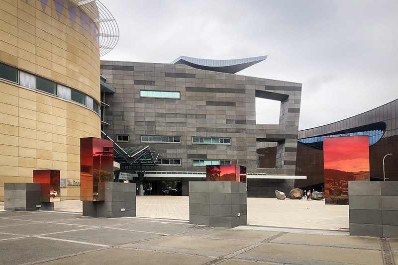 A photo of the forecourt of Te Papa with four concrete blocks lined up. Each of the blocks has a mirrored-glass rectangle at different positions on each.