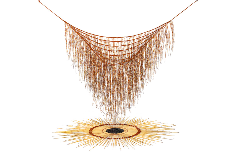 An art work with a woven harakeke (flax) cape is hanging over another woven artwork arranged in a circle on the white ground.