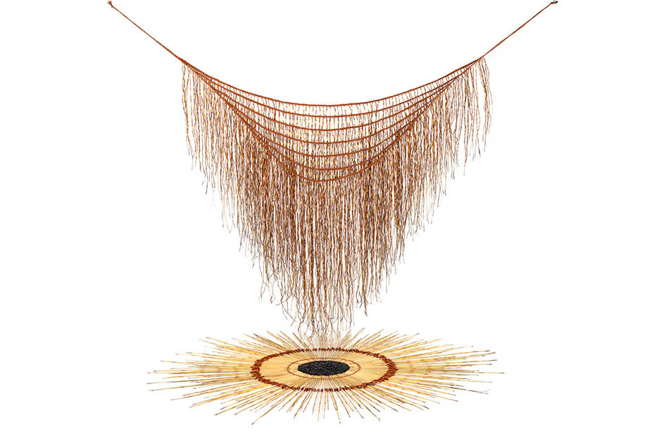 An art work with a woven harakeke (flax) cape is hanging over another woven artwork arranged in a circle on the white ground.