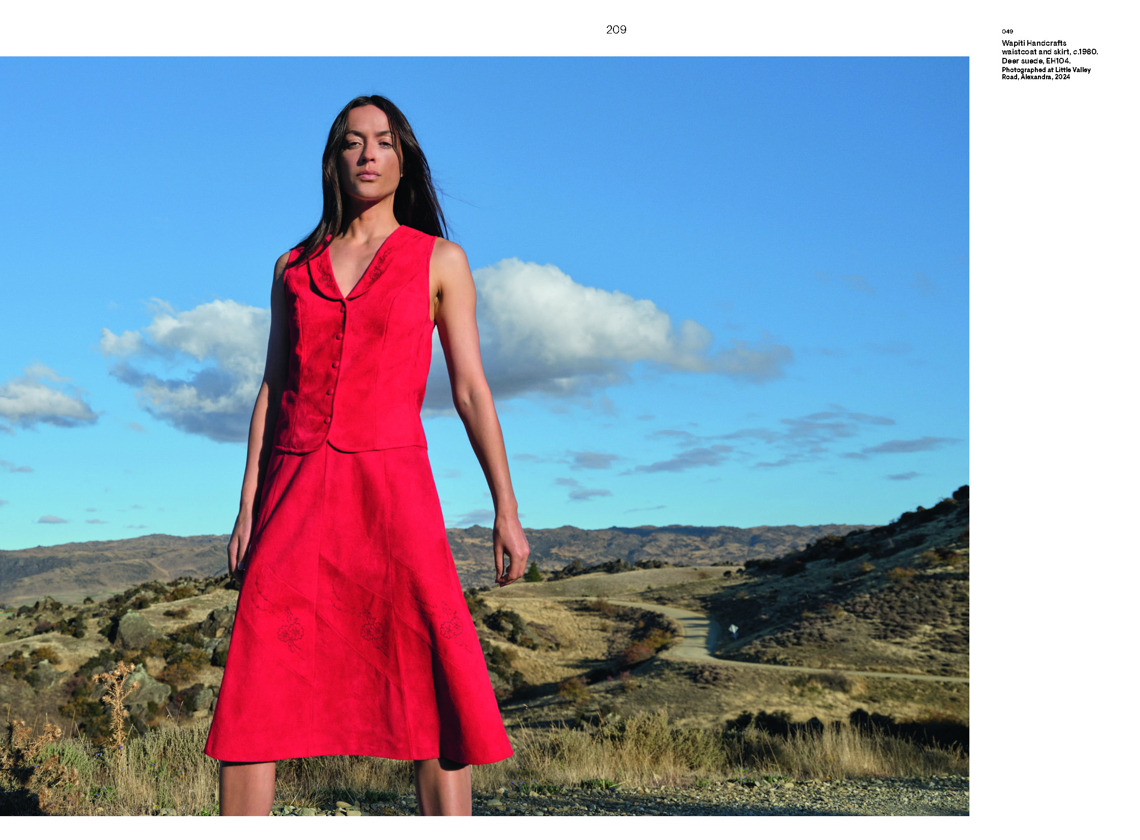 A photo of a woman in a bright red waistcoast and skirt standing in a hilly field.
