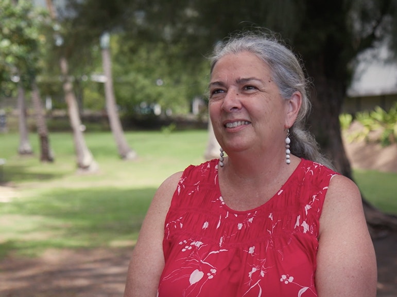 Still from video of Sue Pearson interview. Te Papa A woman with her grey hair tied back in a ponytail and wearing a sleeveless red top is standing on some grass smiling at someone off camera.