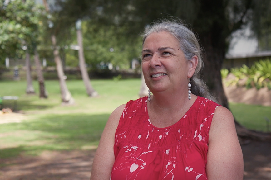 A woman with her grey hair tied back in a ponytail and wearing a sleeveless red top is standing on some grass smiling at someone off camera.