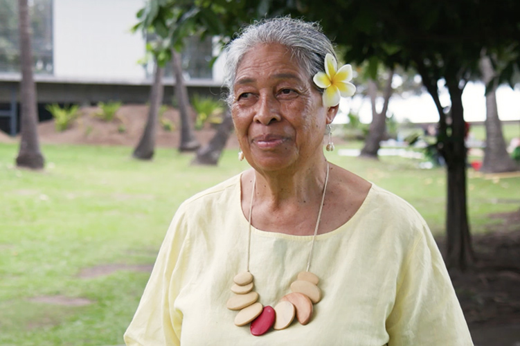 An older woman is standing on grass smiling at someone off camera. She is in a yellow top and is wearing a frangipani flower behind her left ear.