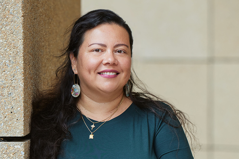 A head and shoulders photo of a woman with long dark hair, and wearing jewellery. She is leaning against a stone pillar and smiling at the camera.