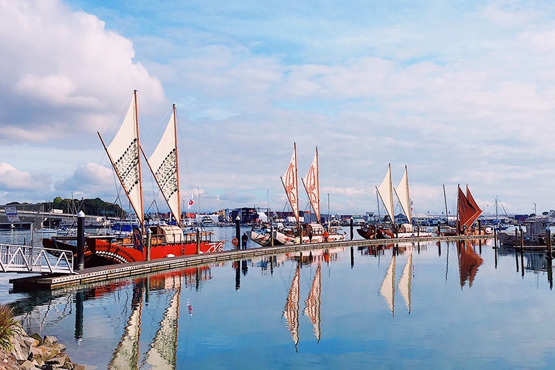 Four double-hulled vessels sailing in a line on very still water in a harbour.