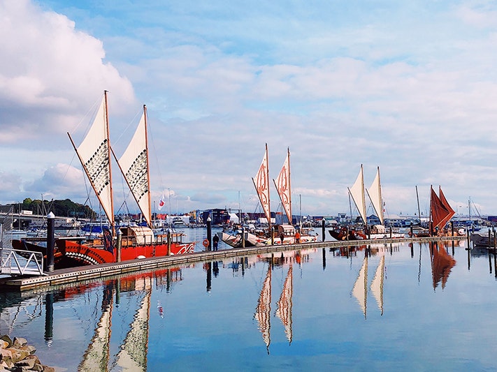 Four waka hourua at Te Hau Komaru, 2021. Photo by Nathan Pettigrew Four double-hulled vessels sailing in a line on very still water in a harbour.