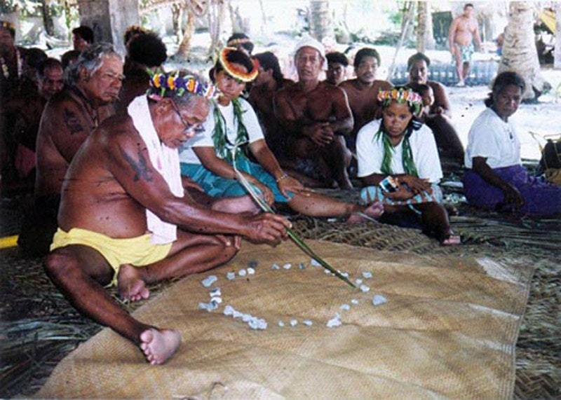 A photo of a man wearing shorts and a towel around his neck sitting on a mat showing others about navigation with a circle of stones that he is pointing at with a stick.