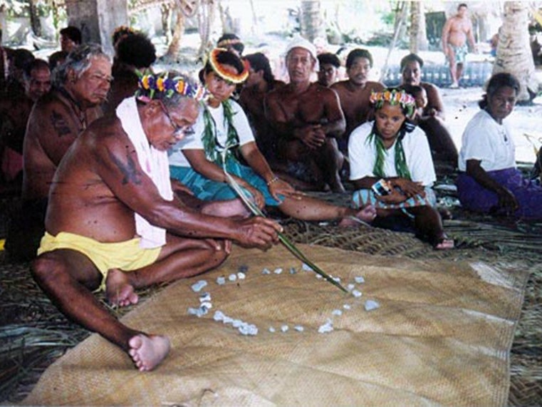 Mau Piailug teaching traditional navigation to a group including members of the Polynesian Voyaging Society. Courtesy of Polynesian Voyaging Society A photo of a man wearing shorts and a towel around his neck sitting on a mat showing others about navigation with a circle of stones that he is pointing at with a stick.