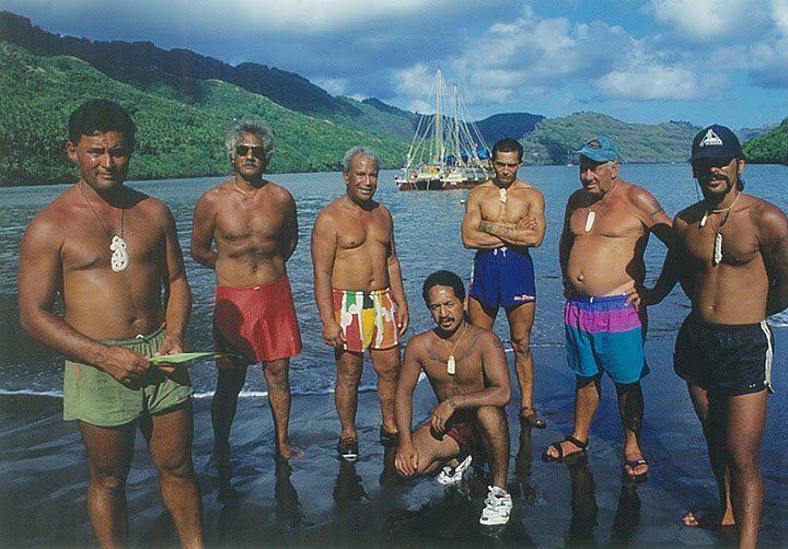 Seven men wearing only shorts are standing on the water's edge. There is a double-hulled vessel in the harbour behind them.