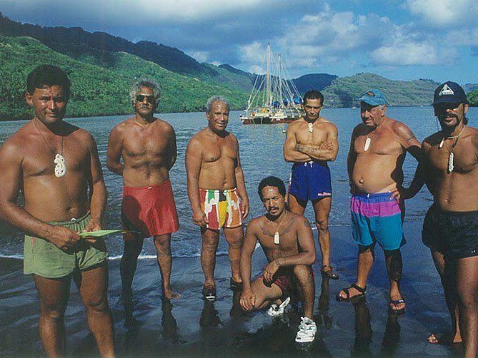 Te Aurere crew members with Nuku Hiva locals, Marquesas, 1995. Courtesy of Te Tai Tokerau Tarai Waka Inc and Jack Thatcher Seven men wearing only shorts are standing on the water's edge. There is a double-hulled vessel in the harbour behind them.